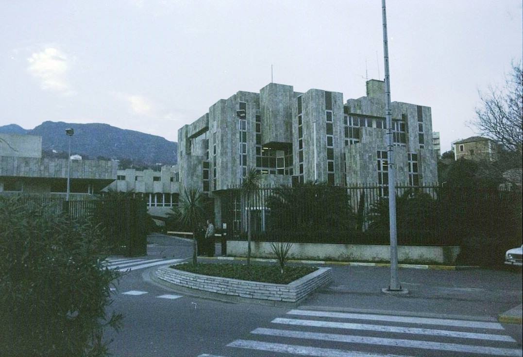 Palazzo di cemento brutale dietro una recinzione, incrocio stradale vuoto e monti grigi