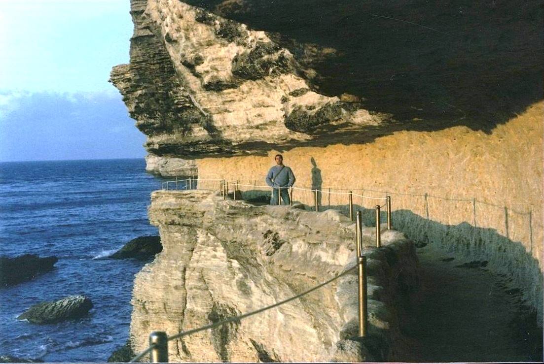 Uomo in maglione blu su sentiero roccioso sopra il mare, sotto parete calcarea