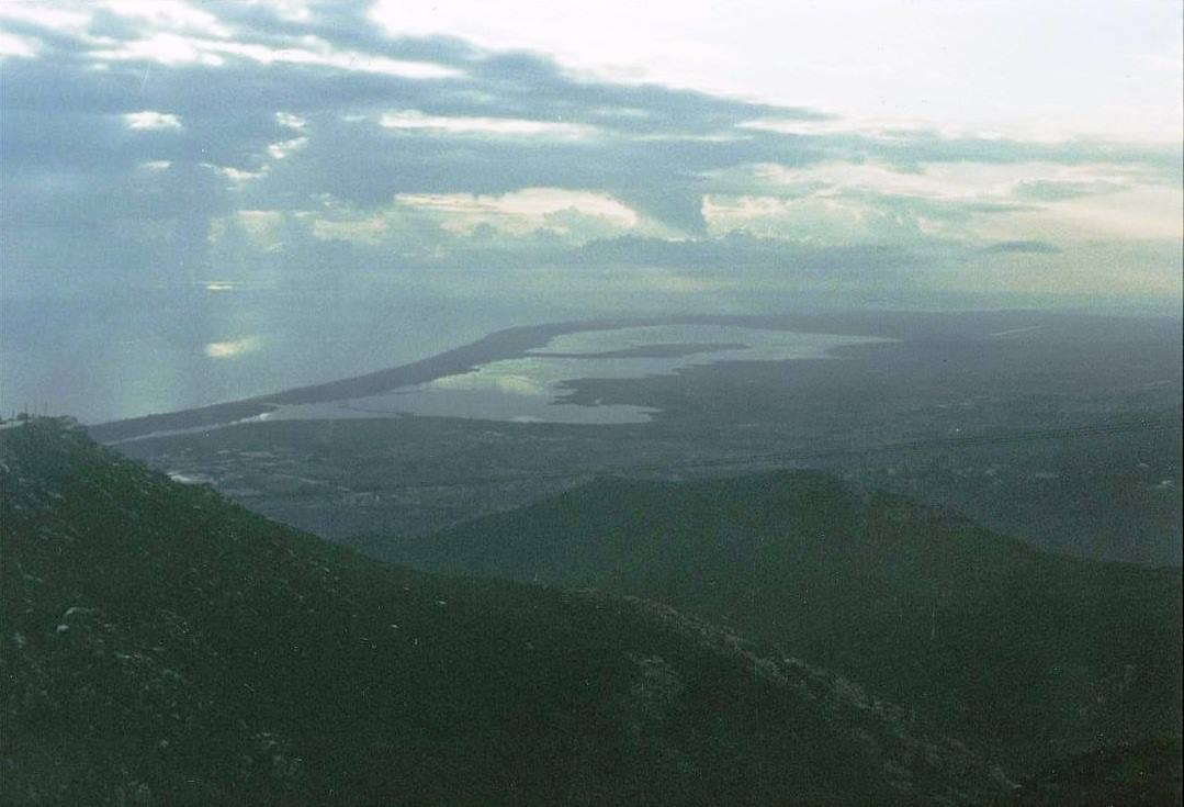 Collina boscosa in primo piano, laguna e costa sotto nuvole con raggi di luce