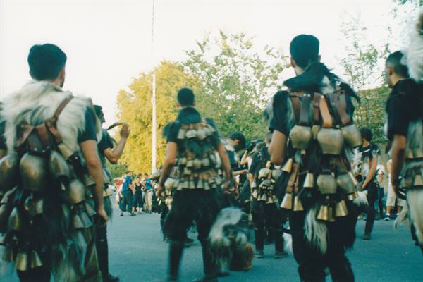 Men in bronze bells and fur costumes dance down the street among the crowd
