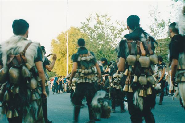 Men in fur costumes with bronze cowbells parade down the street past spectators