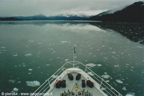Ship bow among floating ice, snow-capped mountains reflected in grey water