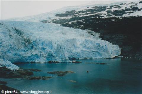 Blue glacier front plunges into dark water among rocks and snow