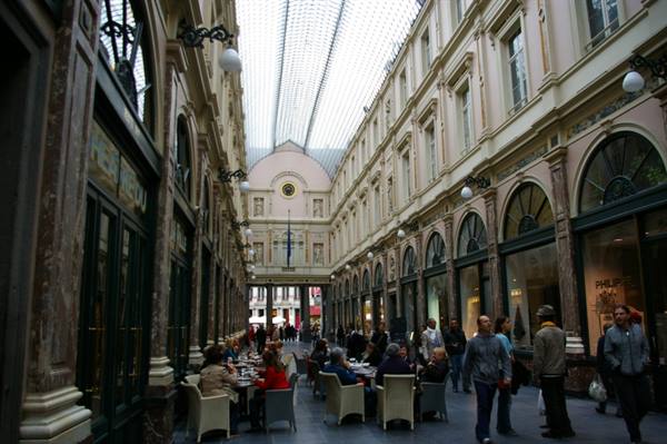 Covered arcade with glass roof, people at café tables and strollers