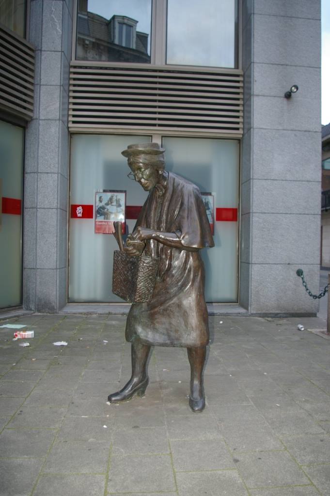 Statua in bronzo di donna anziana col cesto, su marciapiede davanti a un negozio