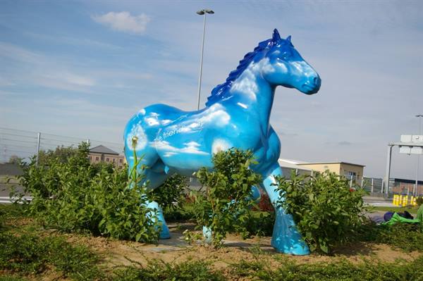 Blue horse statue painted with clouds among green bushes under clear sky