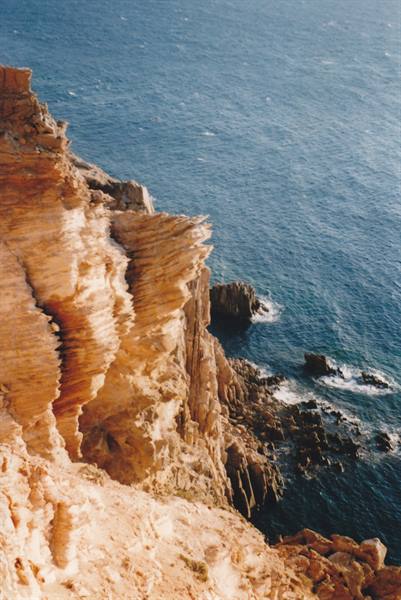Layered orange cliffs plunging into deep blue sea with dark rocks