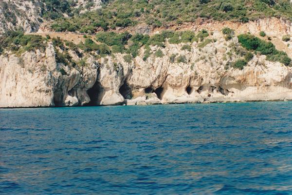 Caves carved in white limestone cliff above blue sea, green shrubs