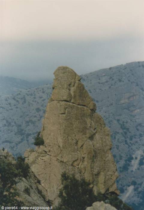 Jagged rock pinnacle rising among Mediterranean shrubs, hazy mountains under overcast sky