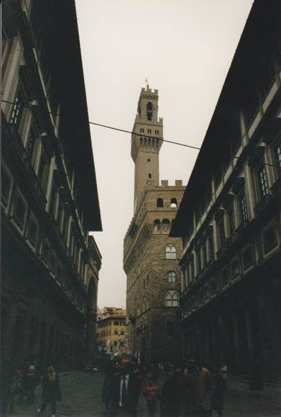 Palazzo Vecchio tower framed by Uffizi corridors, tourists below under grey sky