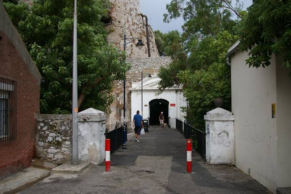 Pedestrians crossing a bridge toward a white gate under stone fortress walls, trees alongside