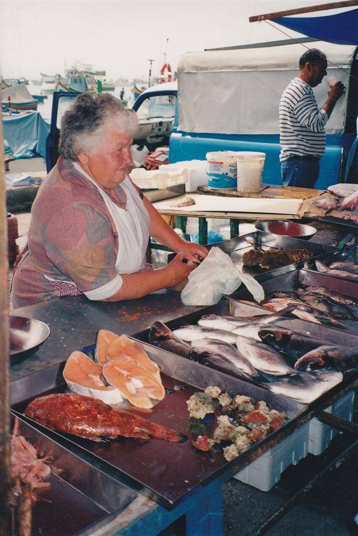 Venditrice al banco del pesce al porto, con banchi metallici e barche sullo sfondo