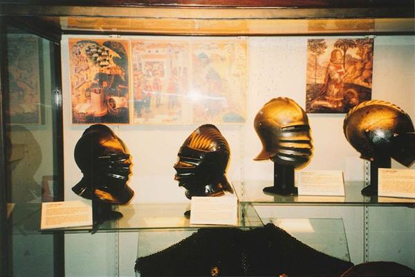 Four medieval steel helmets displayed in museum glass case with frescoes