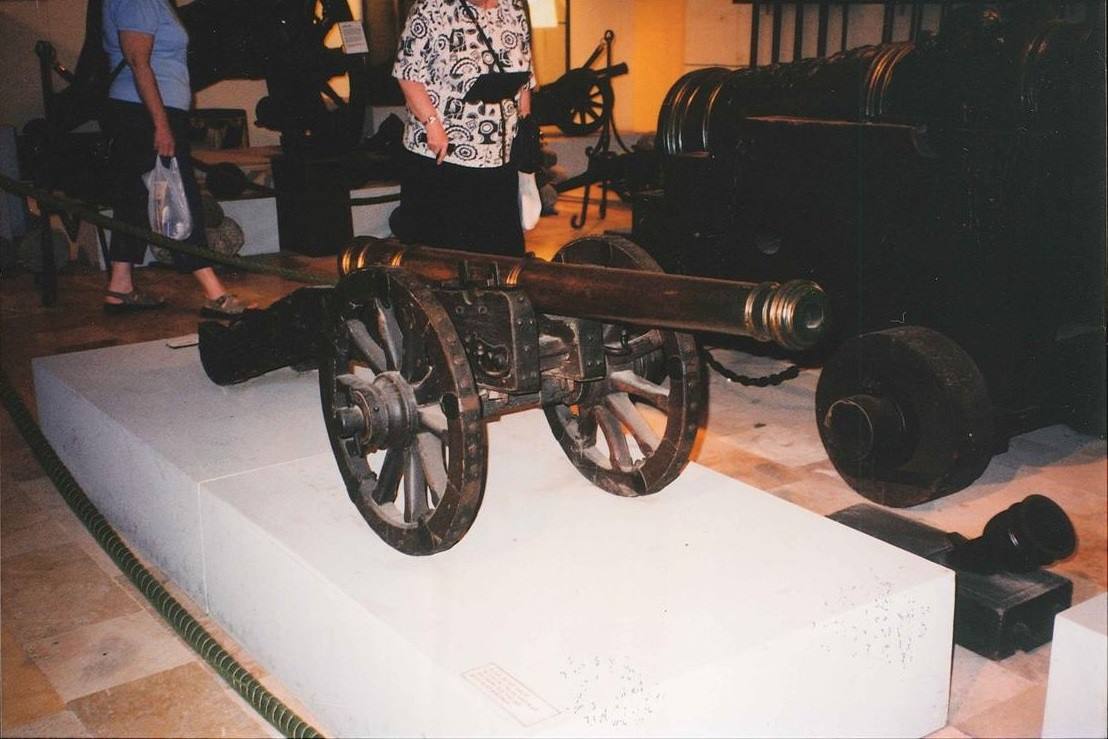 Cannone antico in bronzo con ruote lignee esposto su piedistallo bianco nel museo
