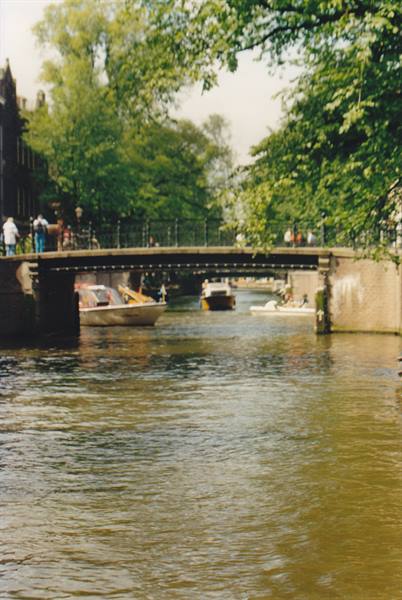 Boats passing under a brick canal bridge in Amsterdam, leafy trees