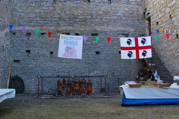 Spit-roasted meat in a stone courtyard with Sardinian flag and colorful bunting