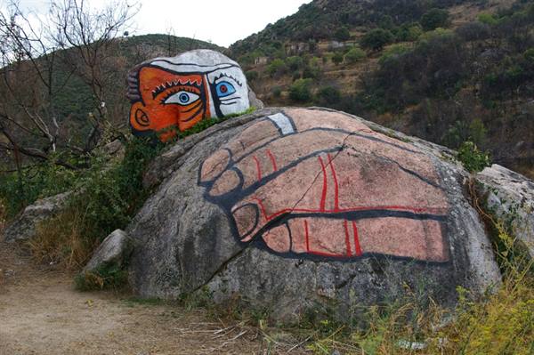 Boulder painted with Picasso-style face and hand among hills and vegetation