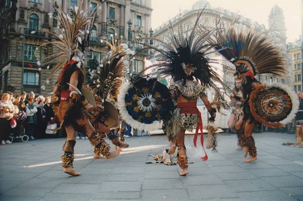 Aztec dancers in feathered costumes and shields dance in a city square