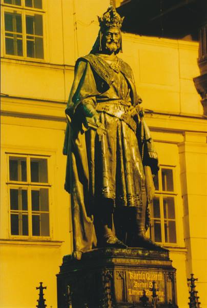 Bronze statue of Emperor Charles IV wearing a crown, in front of a yellow Prague palace