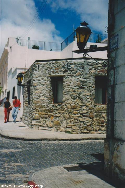Stone house with wrought-iron lanterns on cobblestone street, two people walking, Colonia del Sacramento