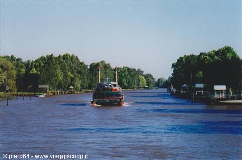 Red steamboat sails a wide river lined with docks and trees