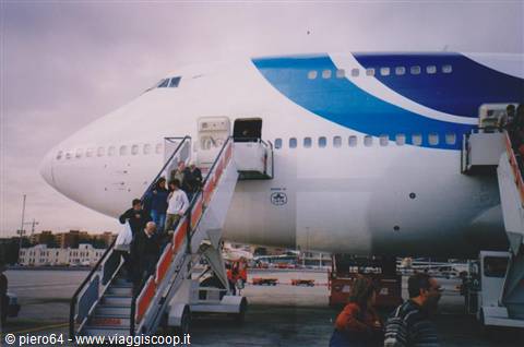 Passengers board a white and blue Boeing 747 via mobile stairs on the apron