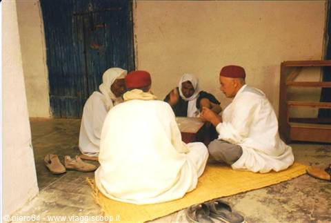 Men in red fezzes and white robes play cards sitting on the floor