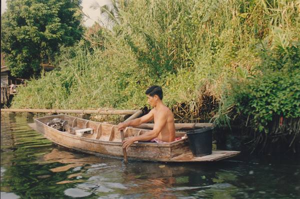 Shirtless man paddles a wooden canoe along a lush green canal