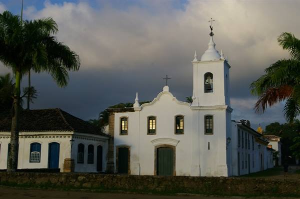 White colonial church with bell tower among palm trees, cloudy sky in Paraty