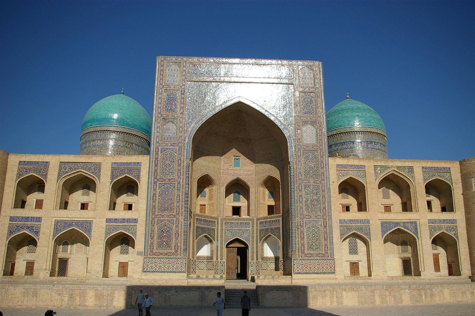 Madrasa con portale a mosaici azzurri e due cupole turchesi, visitatori camminano nella piazza