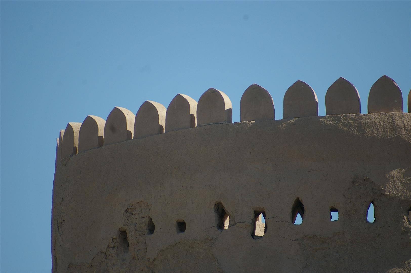 Torre fortificata in fango, merli appuntiti e feritoie contro cielo azzurro