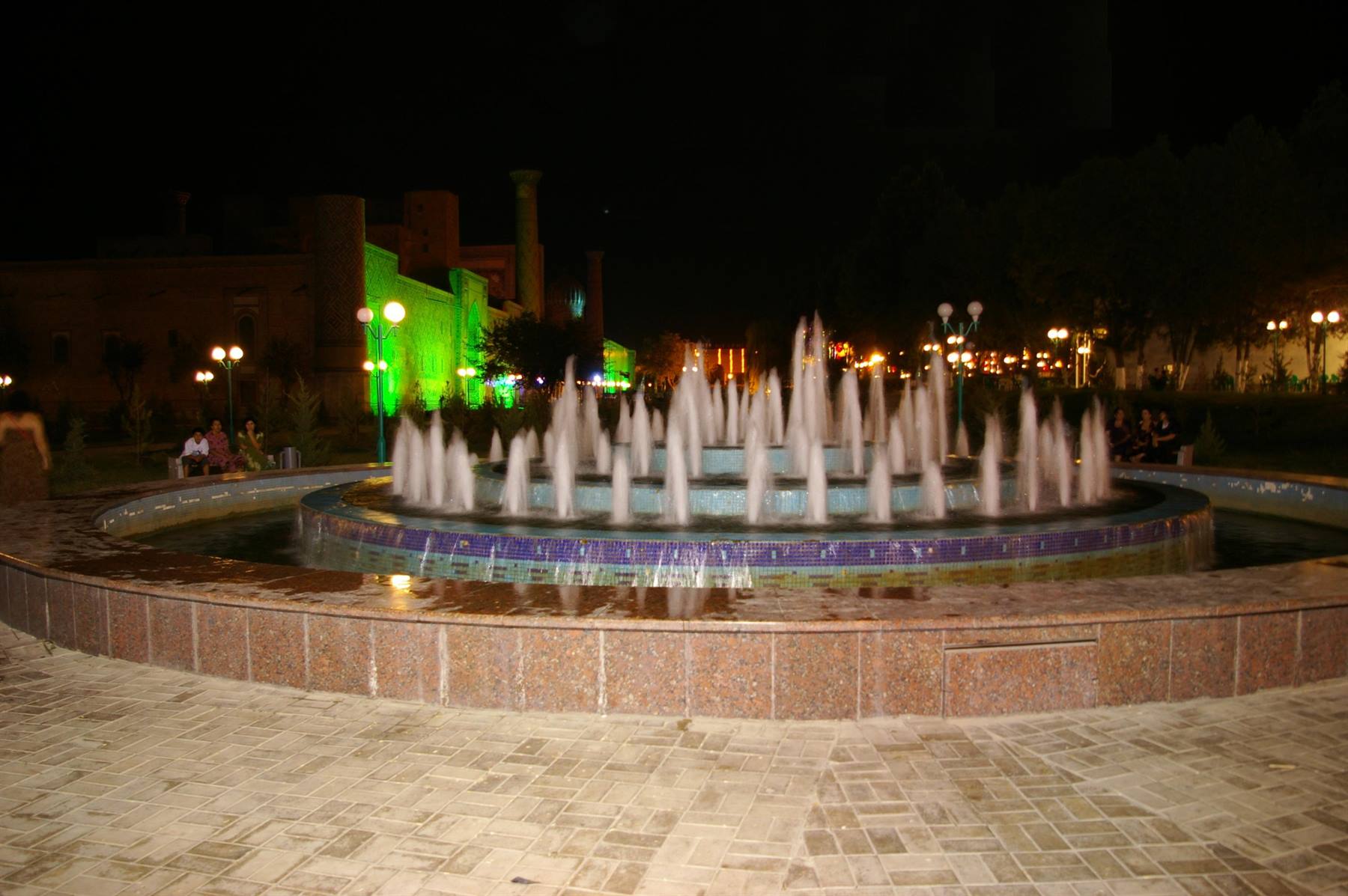 Fontana circolare illuminata di notte, getti d'acqua su pietra, edifici verdi accesi sullo sfondo