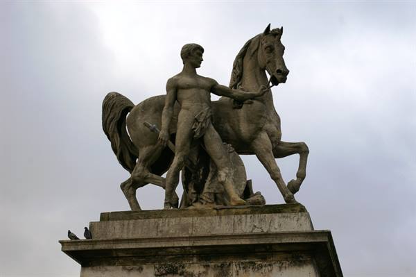 Stone statue of naked man taming a horse, against grey cloudy sky