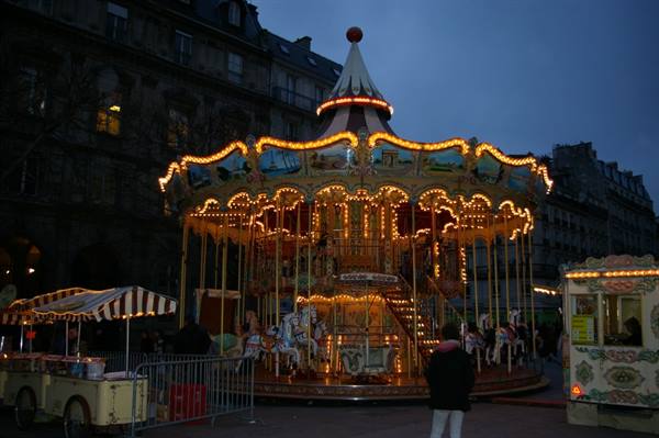 Illuminated carousel at night beside candy stalls, visitors walk past