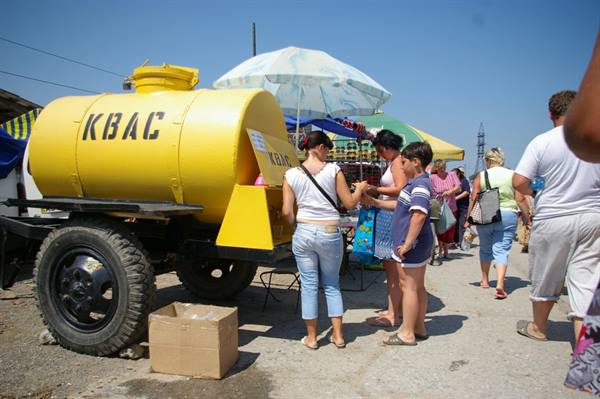 Yellow kvass tank at a street market, people buying drinks under the umbrella