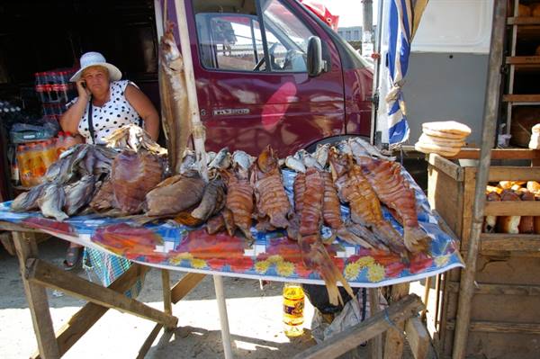 Woman on phone beside a table of dried fish, red van behind