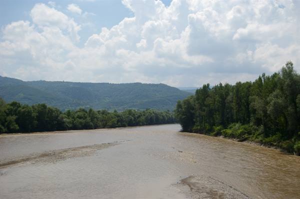 Muddy river between wooded banks and green hills under white clouds