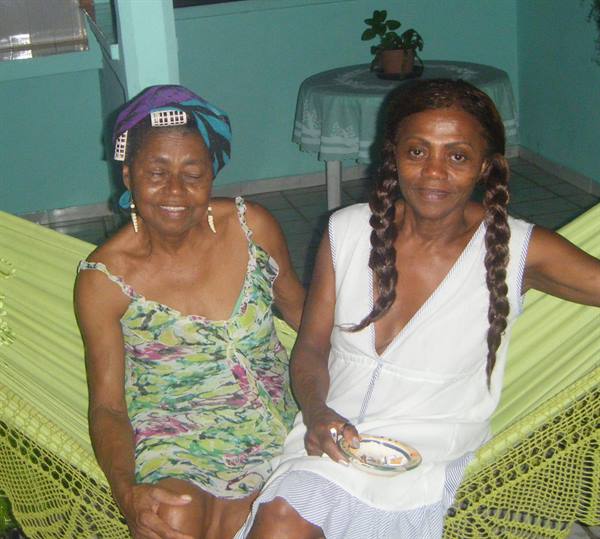 Two smiling women in braid and headwrap on a green hammock, tiled veranda