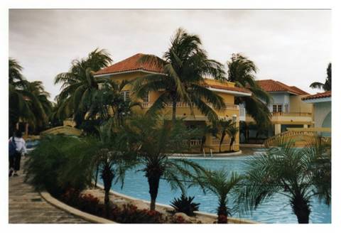 Pool among palms and yellow villas with red roofs, tourists walk under cloudy sky