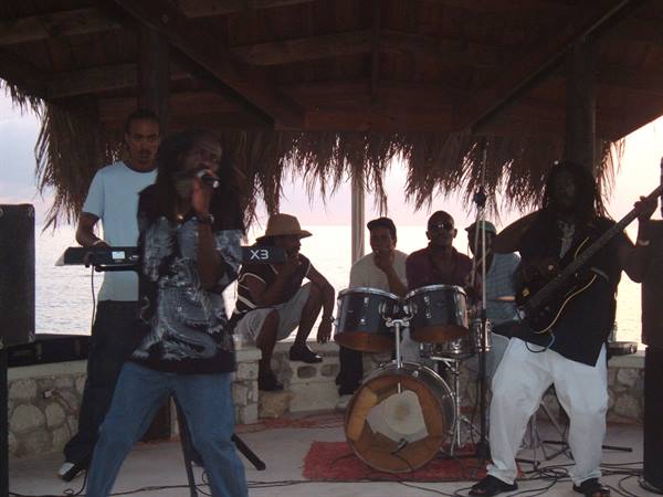Live reggae band under thatched beach hut, drummer, guitarist, singer, audience and sea behind