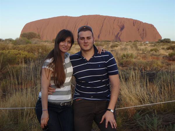 Couple posing in front of Uluru, red rock amid dry grass in morning light