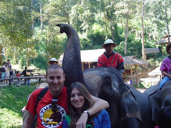 Couple smiles under the elephant's raised trunk, mahout riding behind