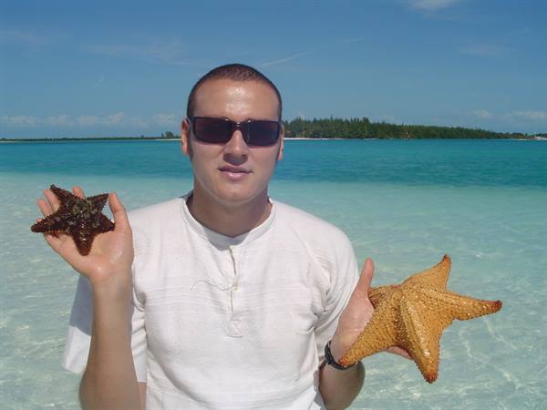 Man in sunglasses holding two starfish in turquoise shallow water, green island behind