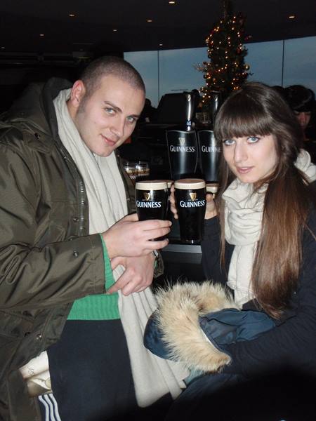 Two young people in scarves raise pints of Guinness near a Christmas tree