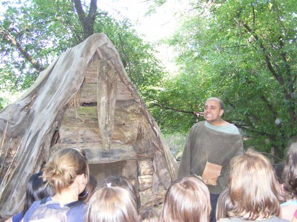 Guide in tunic before a wooden and canvas hut, explaining to a group of visitors