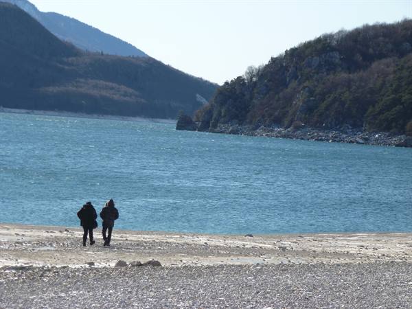 Two people in coats walk on a pebbly shore in front of a blue lake surrounded by wooded mountains