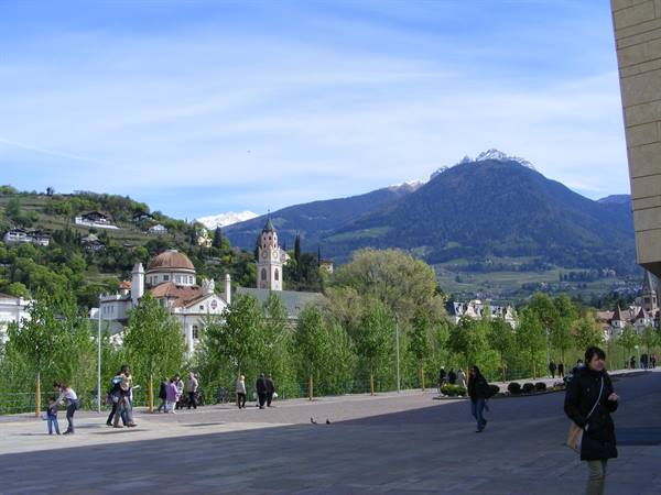People strolling past Merano's churches, snowy peaks under blue sky