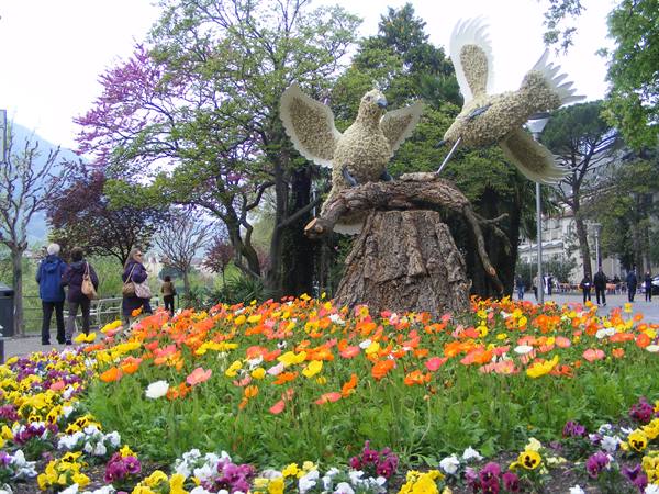 Floral bird sculptures on a tree trunk among colorful flowerbeds and strollers