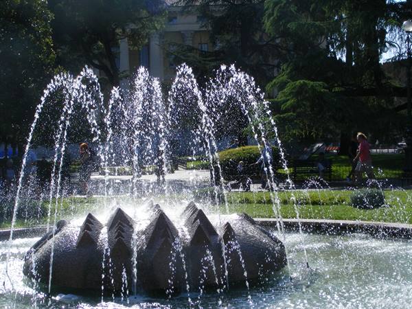 Granite fountain with water jets in a sunny park, people strolling among the trees