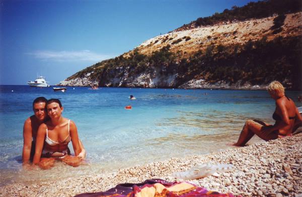 Two girls in the sea on a pebble beach, boat and green hillside